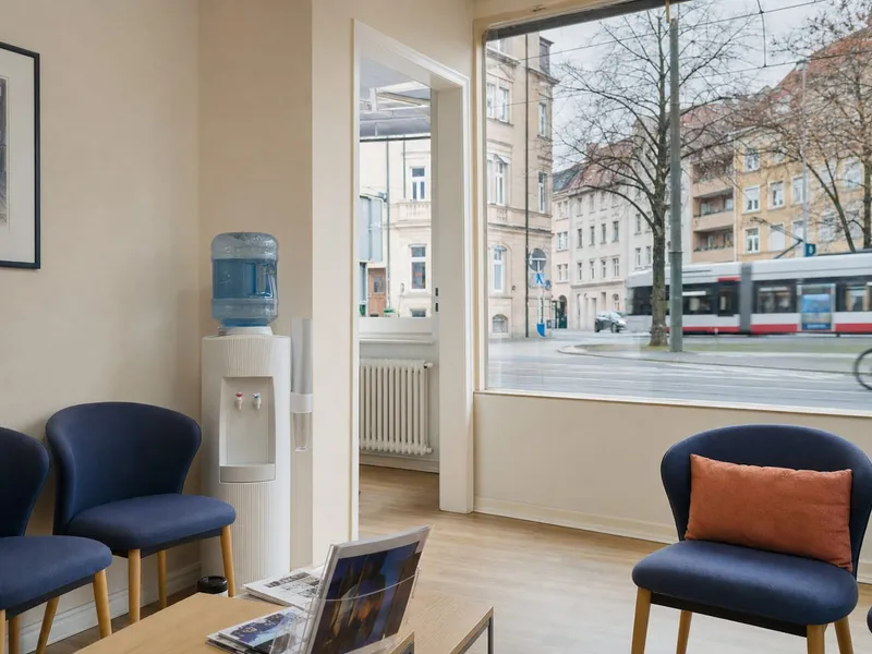 Helles Wartezimmer mit blauen Polsterstühlen, Wasserspender und großem Fenster mit Blick auf eine Straße mit Straßenbahn und Altbauten.